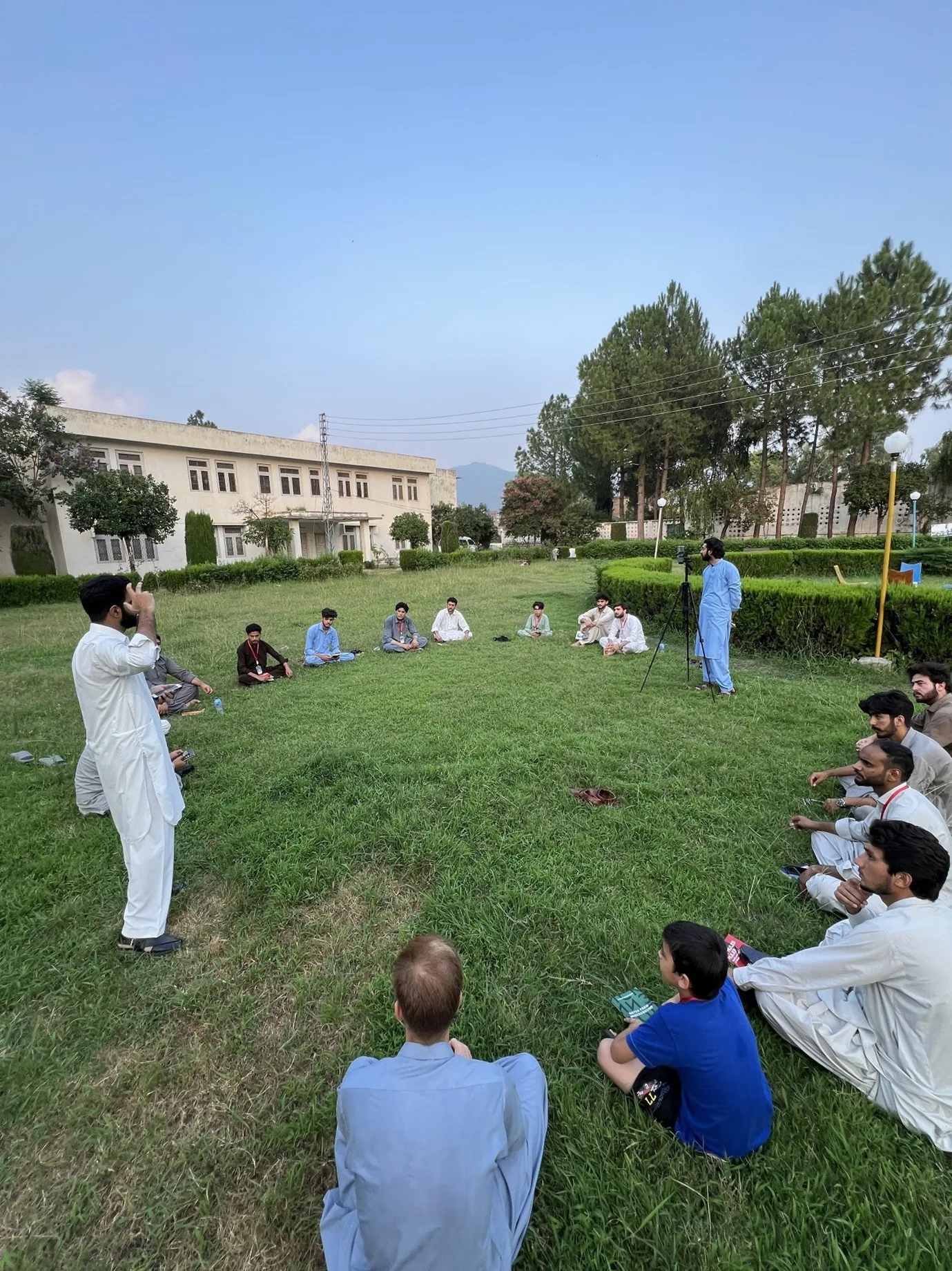 A youth study circle sitting in a circle on grass at dusk.